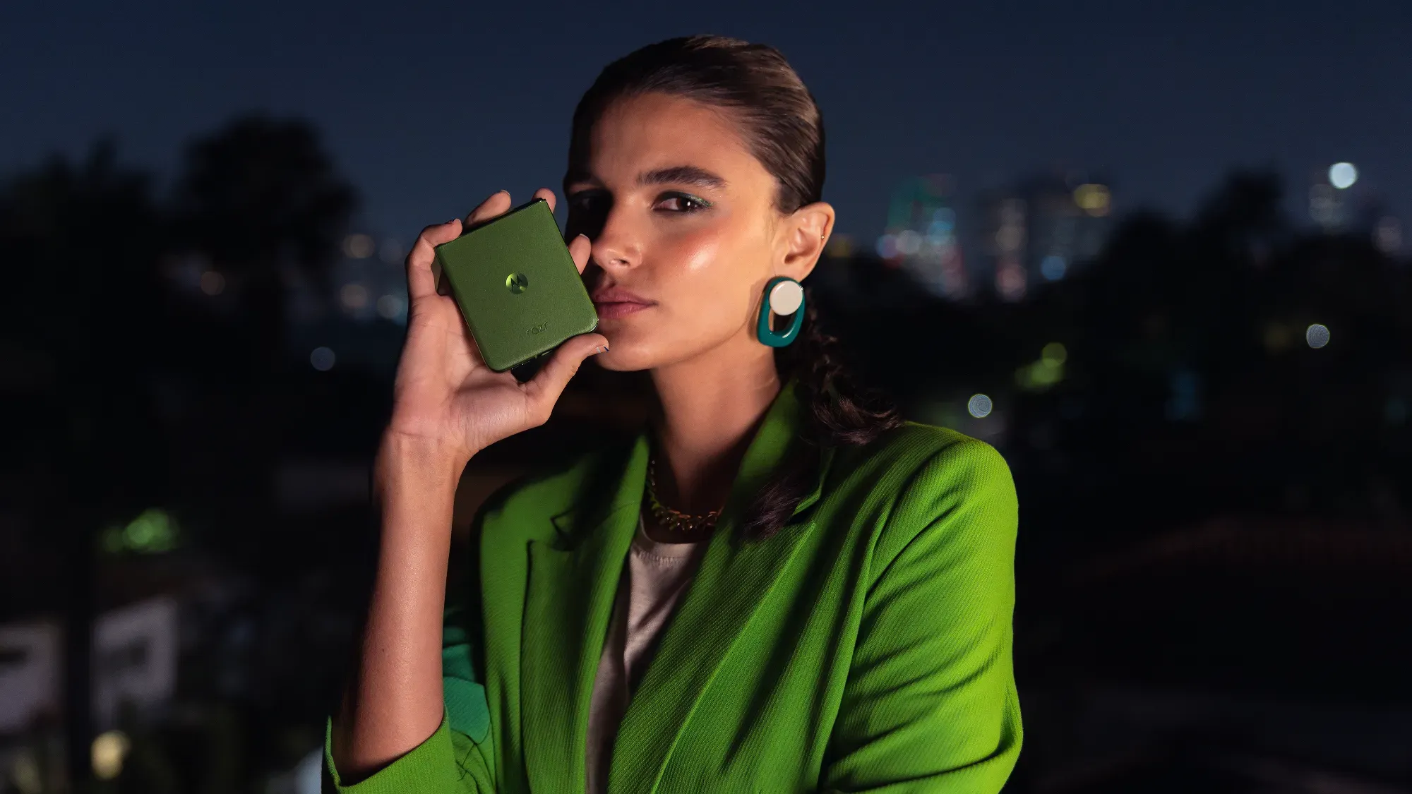 A woman holding a green moto razr  ultra 50 while wearing green earrings and a green jacket. 