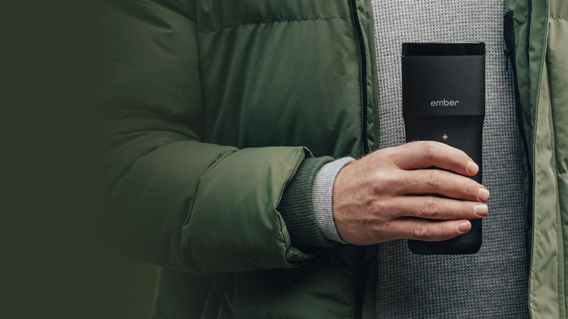 A man's hand in a green jacket and grey jumper holding the Ember Travel Mug 2+