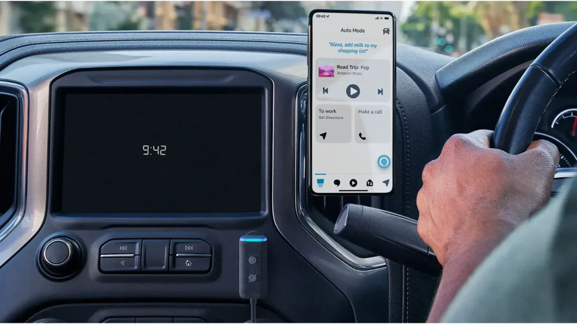 The Amazon Echo Auto in a car near a phone with a man's hand on the steering wheel.