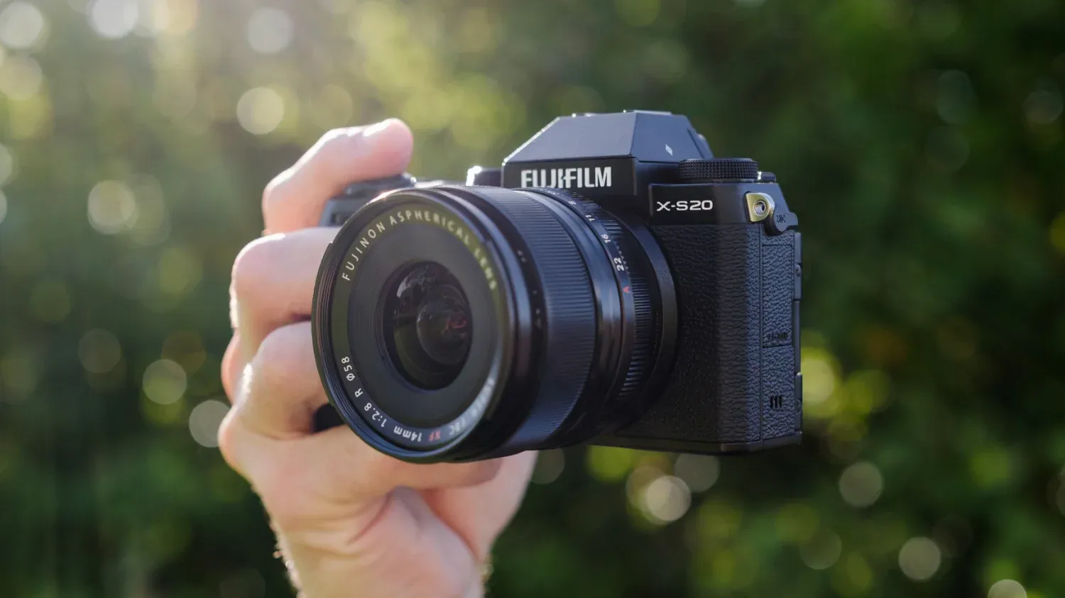 A close up of a hand holding the Fujifilm X-S20, with trees in the background