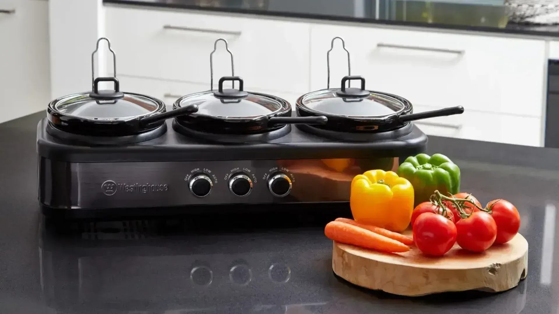 A triple slow cooker behind a chopping board with fruit and vegetables on top