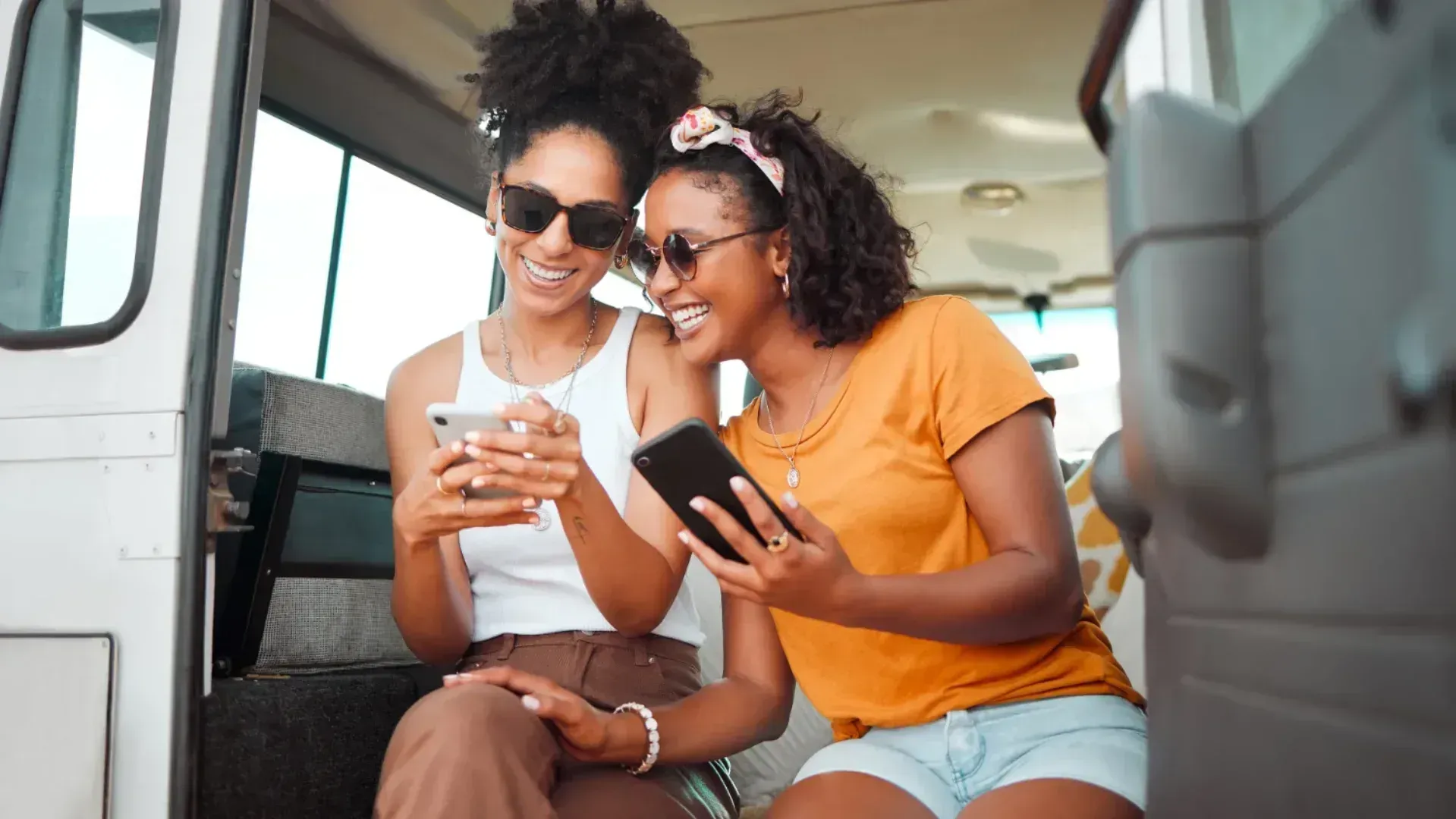 Two young girls looking at their phones from the back of a van, likely enjoying the double data SIM-only deal from Vodafone