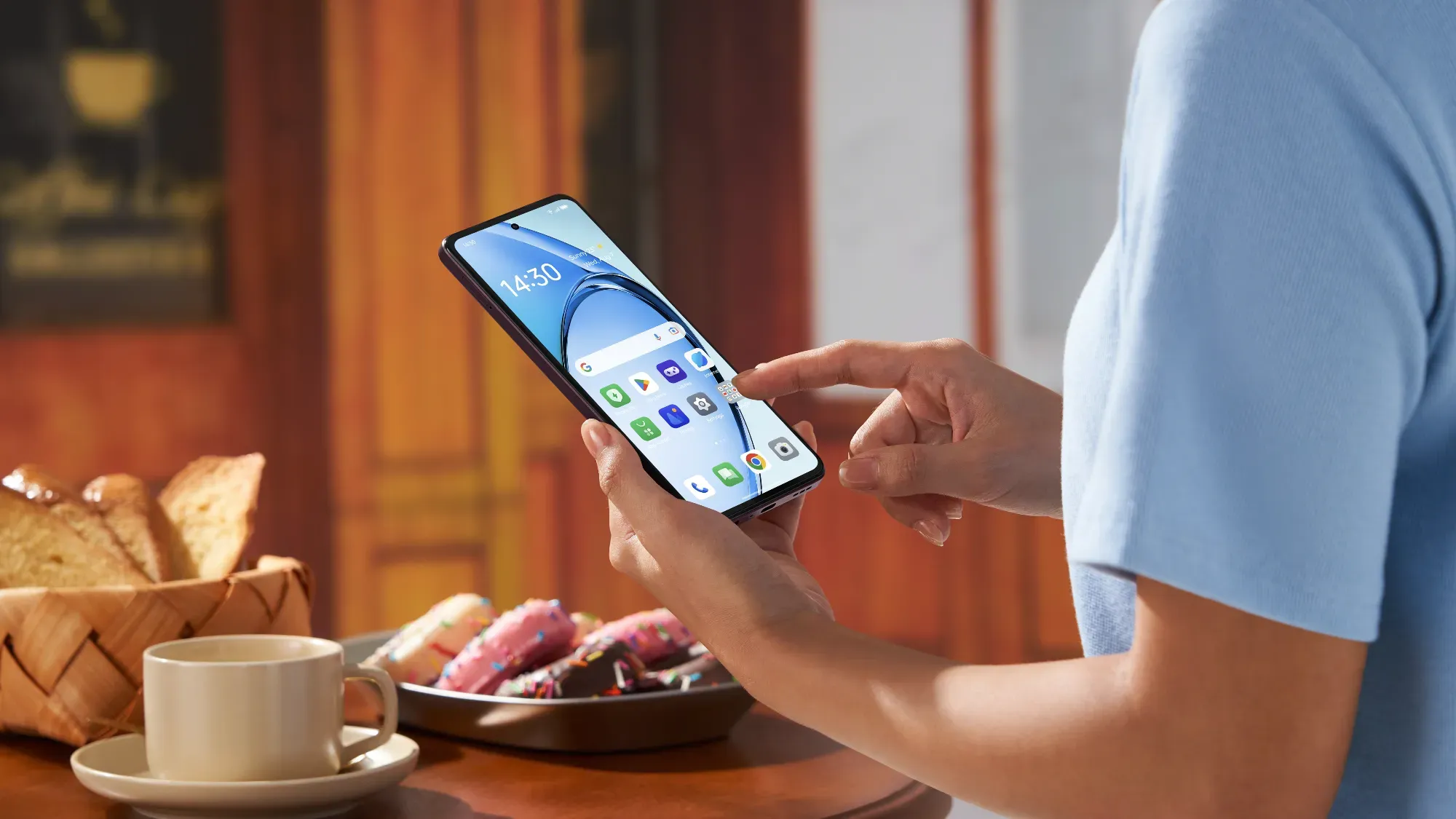 The Oppo A60 5G in hand in a kitchen with coffee and doughnuts.