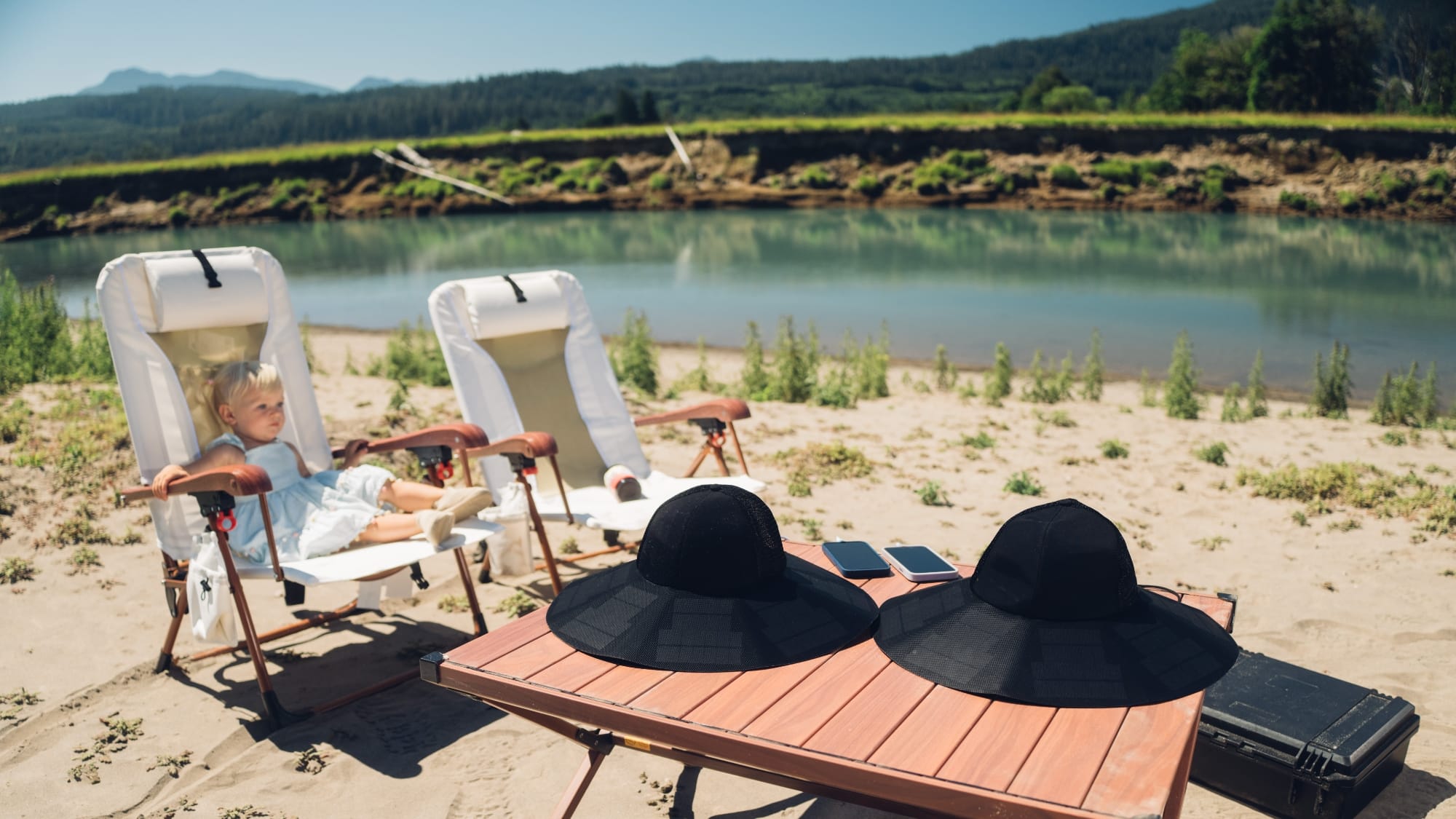 Two EcoFlow Power Hats on a camp table next to a river charging two phones with a child in a chair