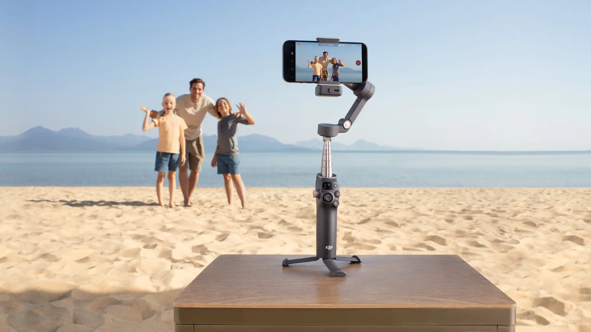 The new Osmo Mobile 7P gimbal on its integrated tripod taking a photo of a man and two kids on a beach