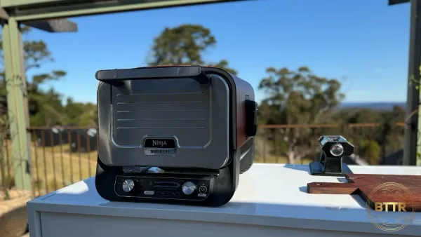 The Ninja Woodfire Outdoor Oven on a benchtop with a pasta roller and a wooden chopping board