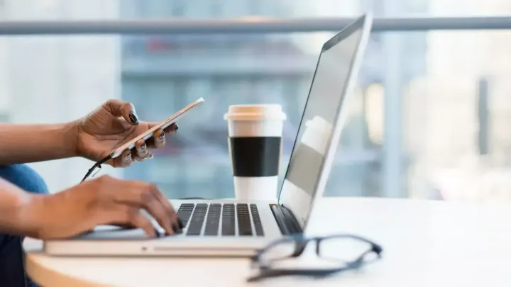 Sideways shot of a laptop with a pair of hands holding a phone and typing and a takeaway coffee cup in the background